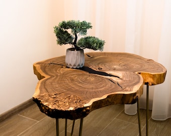 May include: A live-edge wooden side table with a natural, organic shape. The table features a dark, resin-filled crack and is supported by three thin, gold-colored metal legs. A small potted bonsai tree sits on top.