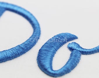 May include: A close-up view of bright blue embroidery thread forming the letters D and a on a white fabric surface. The stitching is dense and raised, creating a textured appearance with a slight sheen on the thread surface.