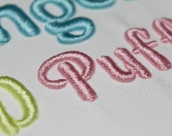 May include: Close-up of embroidered text on white fabric. The word "Puff" is in pink, with other words in light blue and yellow-green. The stitching is thick and textured, creating a 3D effect.