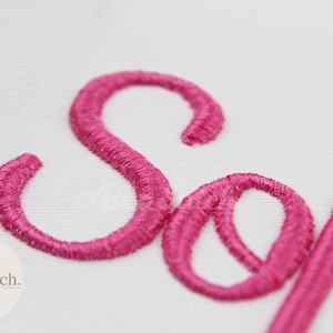 May include: Close-up of the word "Soft" embroidered in bright pink thread on a white fabric. The embroidery is detailed, with each letter clearly defined. The "S" and "o" are in focus.