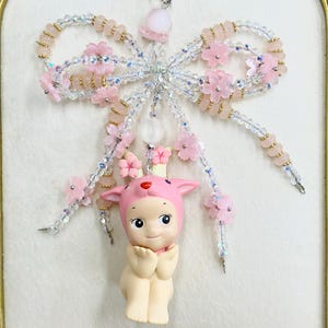 May include: A decorative keychain featuring a large bow constructed from clear iridescent beads and pale pink beads, accented with small pink cherry blossom flowers. A small figurine of a fawn wearing a pink hood hangs from the center of the bow.