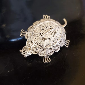 May include: A silver filigree turtle brooch. The brooch features an intricate openwork design with a detailed shell, head, and legs. The silver metal has a bright, reflective finish. The brooch is a decorative accessory.