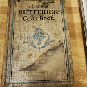 May include: A vintage cookbook titled "The NEW BUTTERICK Cook Book" with a blue and white floral design on the cover. The cover is worn and has some water damage.