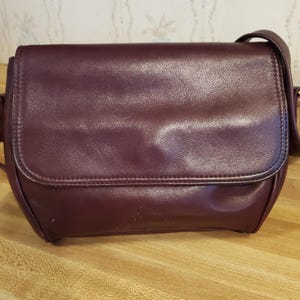 May include: A burgundy leather crossbody bag with a flap closure. The bag has a long, adjustable strap and gold-toned hardware. The bag is sitting on a wooden surface.