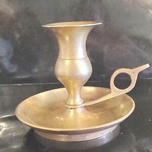 May include: A vintage brass candlestick holder with a wide, shallow dish base and a central cup for the candle. The holder has a handle with a circular opening. The brass has a warm, aged patina.
