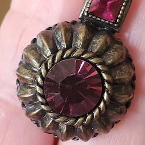 May include: A vintage-style earring with a round, textured, gold-toned metal base and two purple rhinestones. The larger rhinestone is surrounded by a rope-like design.