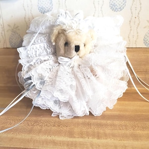 May include: A cream-colored teddy bear with a white lace collar and bow. The bear is surrounded by white lace and ribbons, creating a delicate and vintage aesthetic. The background features a light-colored wallpaper.