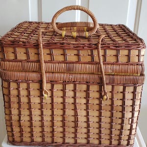 May include: A rectangular picnic basket made of woven wicker in shades of brown and tan. It features a rounded handle on top and a hinged lid. The basket has a woven pattern and a secure closure.