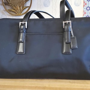 May include: Black tote bag with two top handles and silver buckles. The bag has a rectangular shape with a front pocket and a leather tag attached to one handle. The bag is made of a smooth, matte material.
