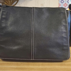 May include: A black leather messenger bag featuring a rectangular flap, a vertical center seam with white contrast stitching, and silver-toned metal hardware attachments for the shoulder strap on both sides of the bag.