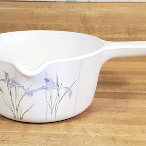 May include: White saucepan with a long handle and a pouring spout. The pan features a floral design of purple irises and green stems. The handle is integrated into the pan's design, and the pan is made of a durable material.