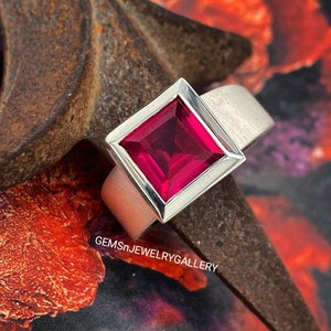 Puede incluir: Un anillo de plata con una piedra preciosa de rubí de forma cuadrada. El anillo tiene un estilo simple y moderno.