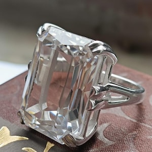 May include: A silver ring featuring a large, rectangular, clear gemstone. The gemstone is faceted, catching the light and creating a sparkling effect. The ring's band has a split design with decorative accents on the sides.