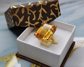 May include: A gold ring featuring a large, rectangular, emerald-cut yellow gemstone set in a four-prong setting. The ring is displayed inside a white square box with a dark brown lid decorated with a gold-colored, branching leaf-like pattern.