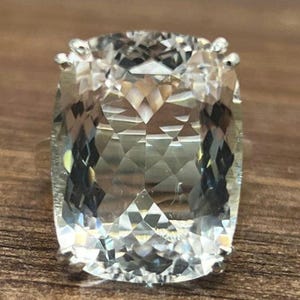 Op de afbeelding: Een grote, heldere, gefacetteerde edelsteenring met een zilveren zetting. De ring ligt op een houten oppervlak.