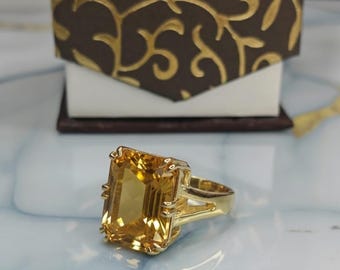 May include: A gold ring featuring a large, rectangular, yellow gemstone. The ring has a gold band and a decorative setting around the gemstone. A brown and gold patterned box is in the background.