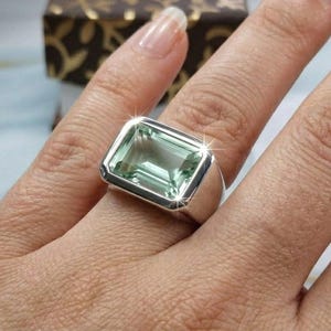 May include: A silver ring featuring a large, rectangular, faceted light green gemstone set in a smooth, polished metal bezel. The stone has a clear, transparent appearance with sharp, clean edges and a prominent, reflective surface.