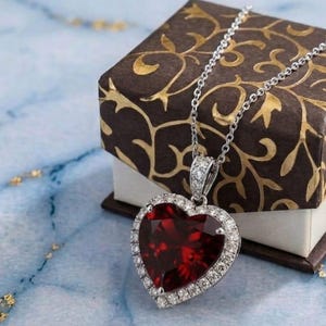 Può includere: Una collana con pendente a forma di cuore con una gemma rossa circondata da un alone di piccole pietre bianche su una montatura metallica. Il pendente pende da una delicata catenina color argento, mostrando una pietra centrale rossa sfaccettata.
