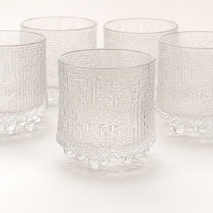 May include: A set of five clear glass tumblers featuring a textured, geometric grid pattern on the exterior walls. Each cylindrical vessel is supported by a series of small, rounded glass spheres arranged along the base, providing a stable foundation.