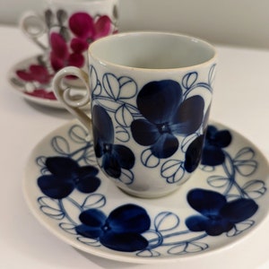May include: White ceramic teacup and saucer set with a blue floral design. The cup has a curved handle and the saucer is round. A second teacup and saucer set is visible in the background, featuring a similar design in pink.