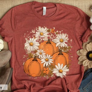 May include: A rust-colored t-shirt with a graphic print of pumpkins and white daisies. The graphic has a distressed, vintage look.