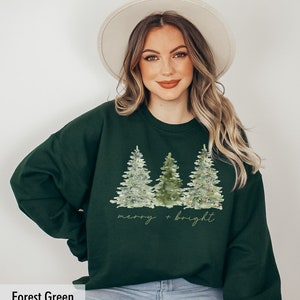 May include: Forest green sweatshirt featuring a watercolour design of three Christmas trees with lights and the text "merry + bright".
