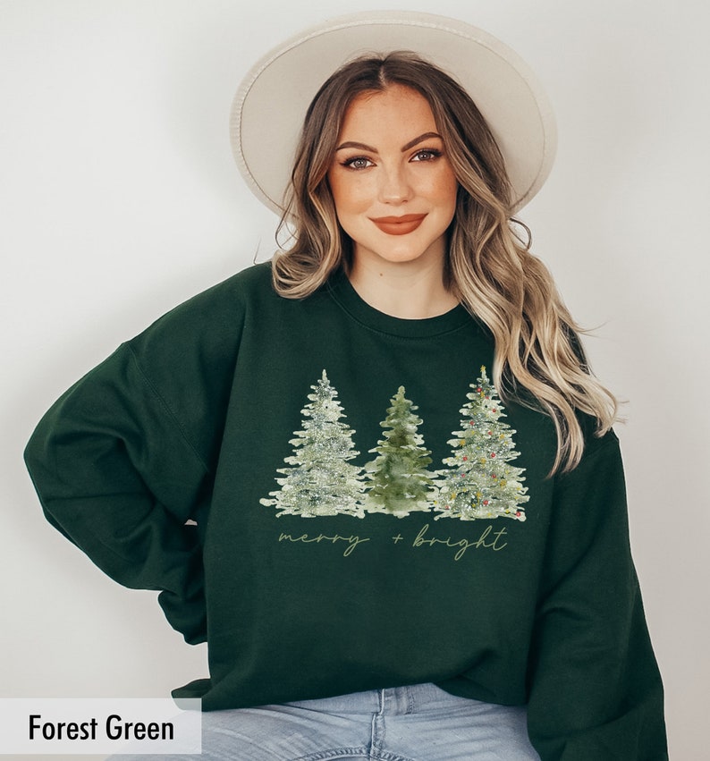 May include: Forest green sweatshirt featuring a watercolour design of three Christmas trees with lights and the text "merry + bright".