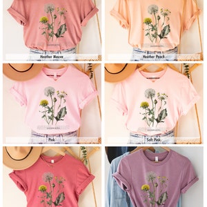 May include: Five different colored t-shirts with a dandelion graphic and the word "DANDELIONS" printed on the front. The colors are heather mauve, heather peach, pink, soft pink, and heather orchid.