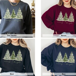 May include: A Christmas sweatshirt featuring a watercolour design of three pine trees with lights and the text "merry + bright". The sweatshirt is available in four colours: dark heather, maroon, black, and navy.