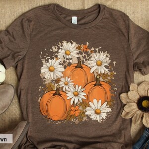May include: A brown t-shirt with a graphic print of pumpkins and daisies. The graphic is in a distressed style with a white background. The text "Heather Brown" is visible at the bottom of the image.