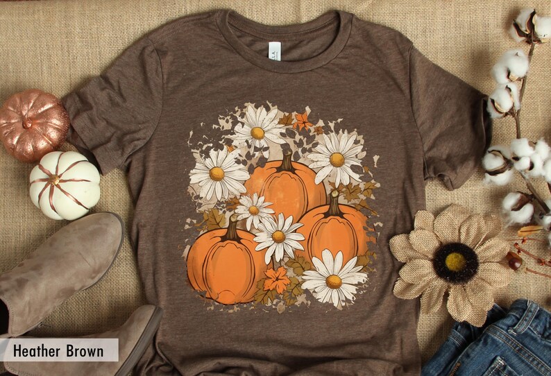 May include: A brown t-shirt with a graphic print of pumpkins and daisies. The graphic is in a distressed style with a white background. The text "Heather Brown" is visible at the bottom of the image.