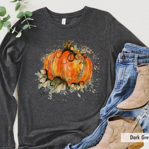 May include: A dark gray heather long-sleeve shirt with a watercolor-style pumpkin design. The pumpkin is orange with green leaves and vines. The design has a splattered paint effect.
