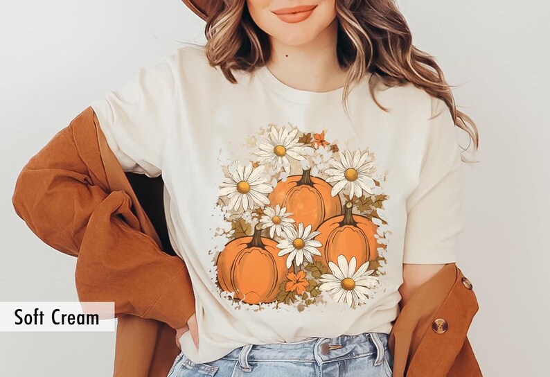 May include: A soft cream t-shirt with a graphic print of orange pumpkins and white daisies. The graphic is centered on the shirt.