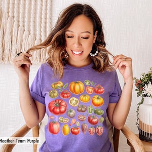 May include: A purple t-shirt with a colorful illustration of various tomatoes, including whole tomatoes, tomato slices, and tomato stems. The text "Heather Team Purple" is visible at the bottom of the image.