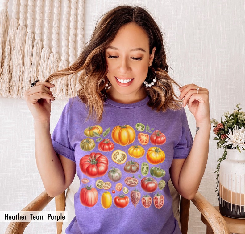 May include: A purple t-shirt with a colorful illustration of various tomatoes, including whole tomatoes, tomato slices, and tomato stems. The text "Heather Team Purple" is visible at the bottom of the image.