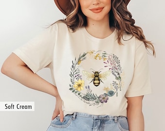 May include: A soft cream t-shirt with a floral wreath and a bee in the center. The wreath is made of yellow, purple, and blue flowers and green leaves.