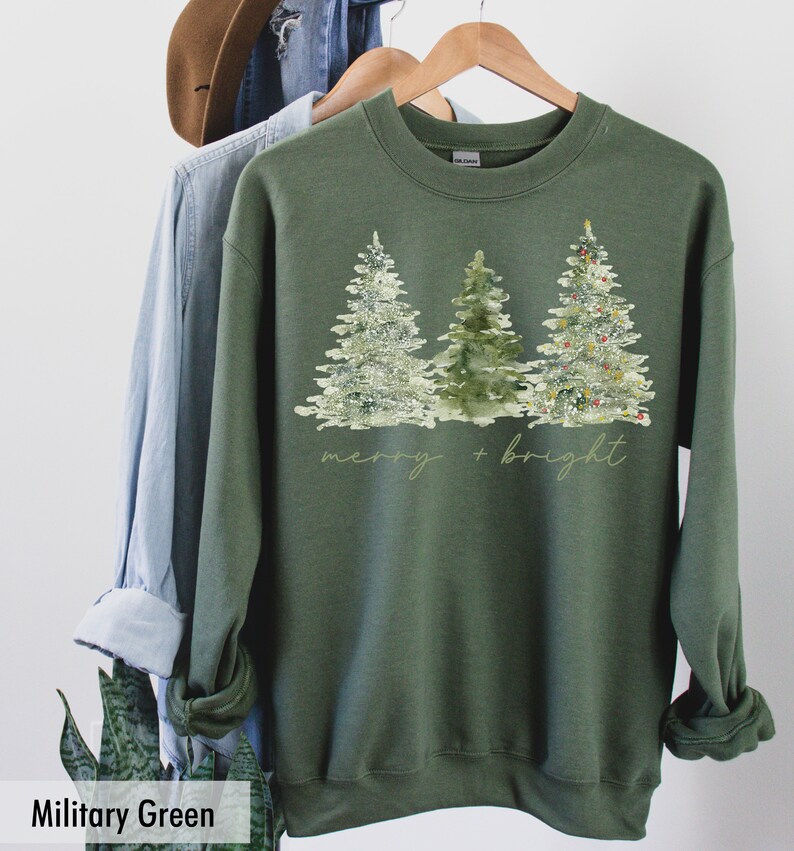 May include: A green sweatshirt featuring a watercolour design of three Christmas trees and the text "merry + bright".