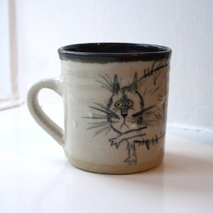 May include: A handmade ceramic mug with a whimsical cat illustration. The mug features a cream-colored body with a dark rim and handle. The cat is drawn in black with yellow eyes, adding a playful touch to this unique piece. A perfect gift for cat lovers.