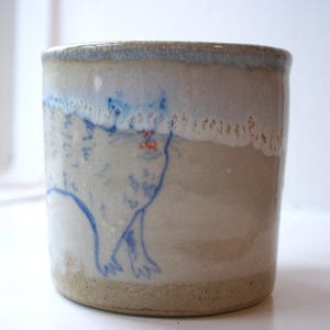 May include: A handmade ceramic mug with a whimsical cat illustration. The mug has a light beige base with a blue cat drawing and a red mouth. The rim has a blue and white glaze. A unique, artistic piece for beverages.