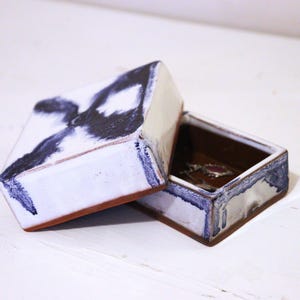 May include: A white ceramic box with a blue and black abstract design. The box has a lid and is open to reveal a brown interior.