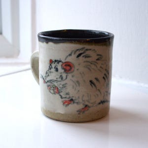 Pode incluir: Caneca de cerâmica com ilustração de rato desenhada à mão. A caneca tem um corpo creme com uma borda e alça marrom escuro. O rato é retratado em cinza, vermelho e preto, segurando um pequeno objeto. Adequada para bebidas quentes ou frias.