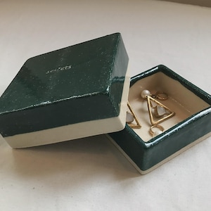 May include: A dark green ceramic jewelry box with a white interior and a white trim around the lid. The box is open and contains a pair of gold triangle earrings with white pearl accents.