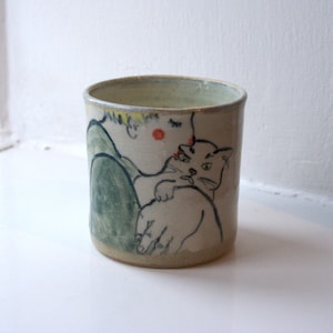 Peut inclure: Un mug cylindrique en céramique avec une illustration dessinée à la main d'une personne embrassant un chat. La personne a les cheveux jaunes, une joue rouge et porte un vêtement vert. Le chat est gris avec des yeux verts. Le mug est beige avec un bord vert clair.