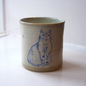 May include: A cylindrical ceramic mug featuring a blue hand-drawn cat illustration. The cat has a red nose and is sitting upright. The mug is light beige with a textured surface and a light blue rim.