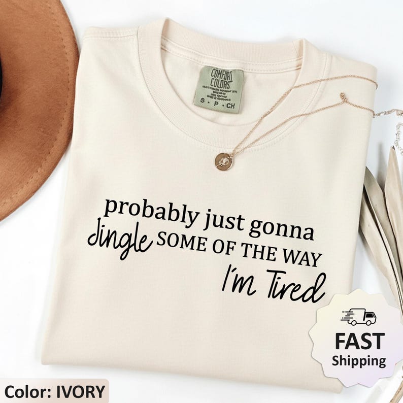 May include: Ivory-colored t-shirt with the text "probably just gonna jingle some of the way I'm Tired" in black script. A gold necklace with a pendant is draped on the shirt. The shirt has a Comfort Colors tag.