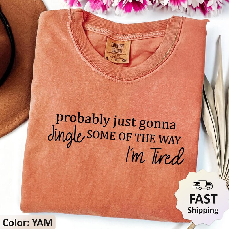 May include: A yam-colored t-shirt with the text "probably just gonna jingle some of the way I'm Tired" in black. The shirt has a crew neck and a Comfort Colors tag. The image also includes a brown hat and flowers.