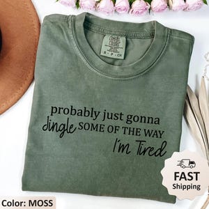 May include: A moss green t-shirt with the text "probably just gonna dingle some of the way I'm Tired" in black. The shirt has a crew neck and short sleeves. The tag reads "Comfort Colors".
