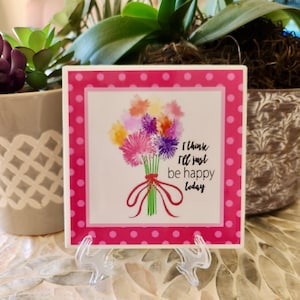 May include: A pink and white ceramic tile with a pink polka dot border. The tile features a watercolor illustration of a bouquet of colorful flowers with a red ribbon and the text "I think I'll just be happy today".