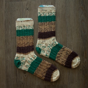 Può includere: Due paia di calzini a righe marroni, verdi e bianchi. I calzini hanno un motivo a maglia.