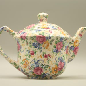 May include: A floral-patterned teapot with a lid and handle. The teapot features a cream-colored base adorned with pink, yellow, and blue flowers. The spout and handle curve gracefully, and the lid has a small knob.